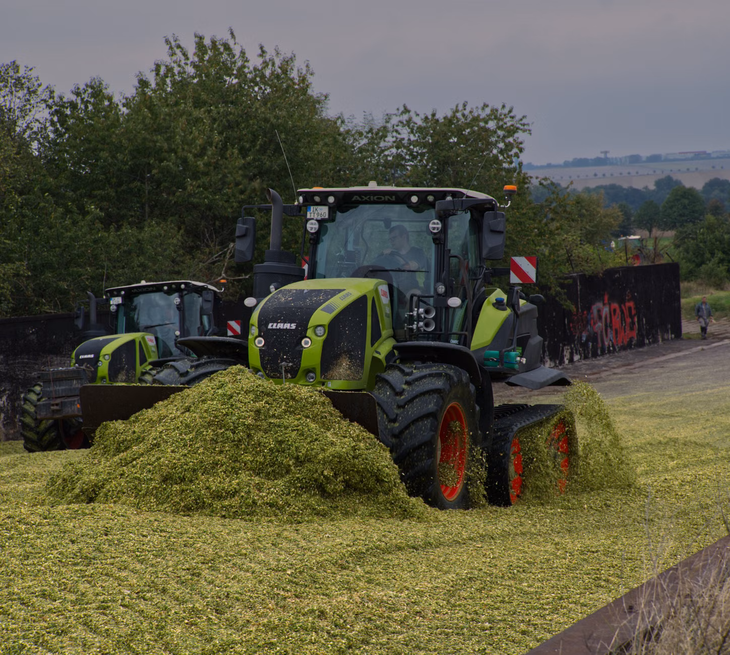 Egy modern CLAAS traktor silóz egy nyílt mezőn, vidéki háttérrel.