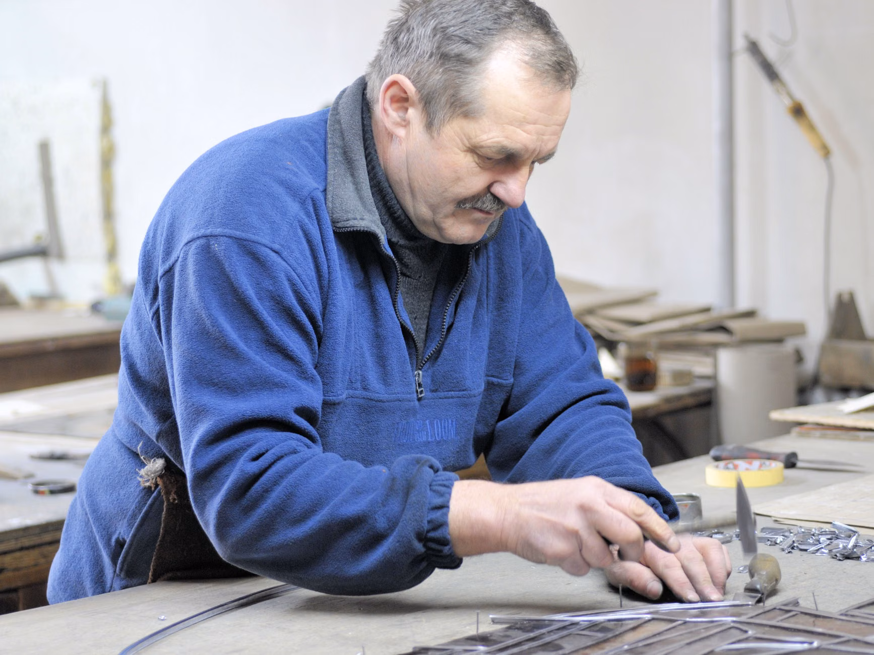 Egy idős férfi, aki fémalkatrészek készítésére összpontosít egy műhely környezetében.