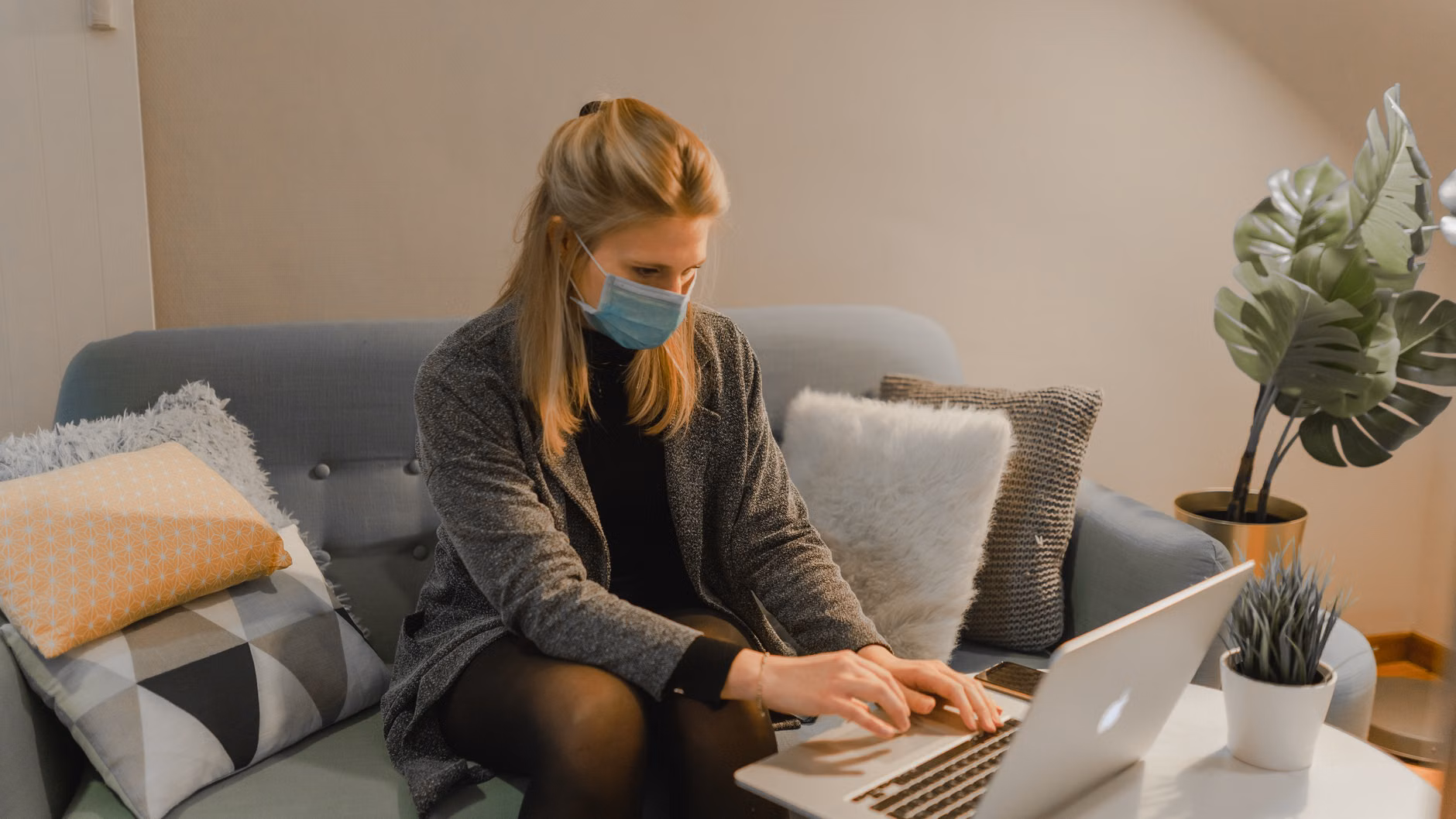 Eine Frau sitzt auf einem Sofa, trägt eine Mundschutzmaske und arbeitet drinnen an einem Laptop.