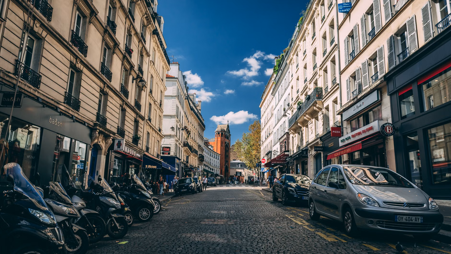 Scène de rue pittoresque à Paris avec une architecture classique, des motos garées et des voitures.