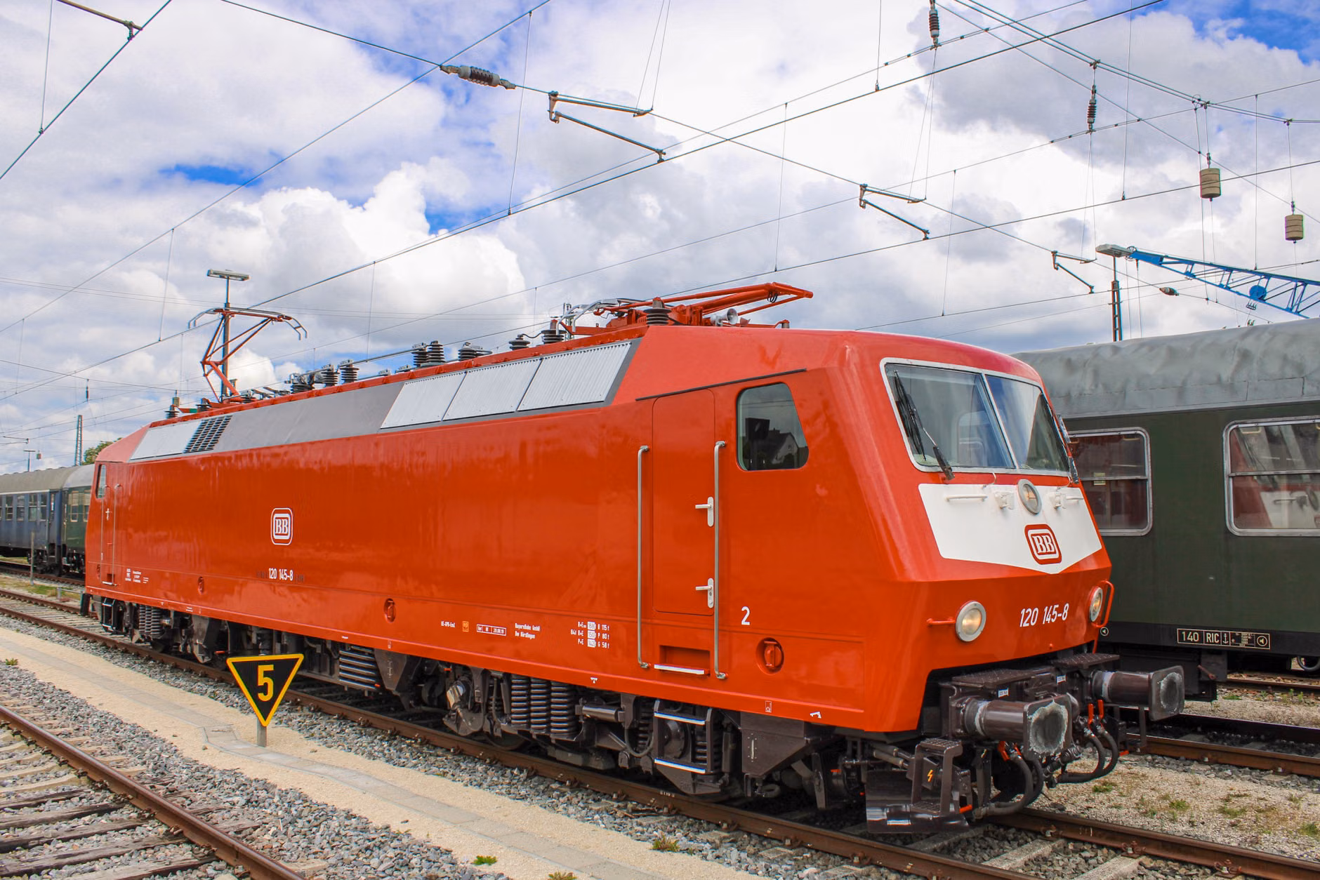 A vivid red locomotive on tracks at a railway yard under a cloudy sky.