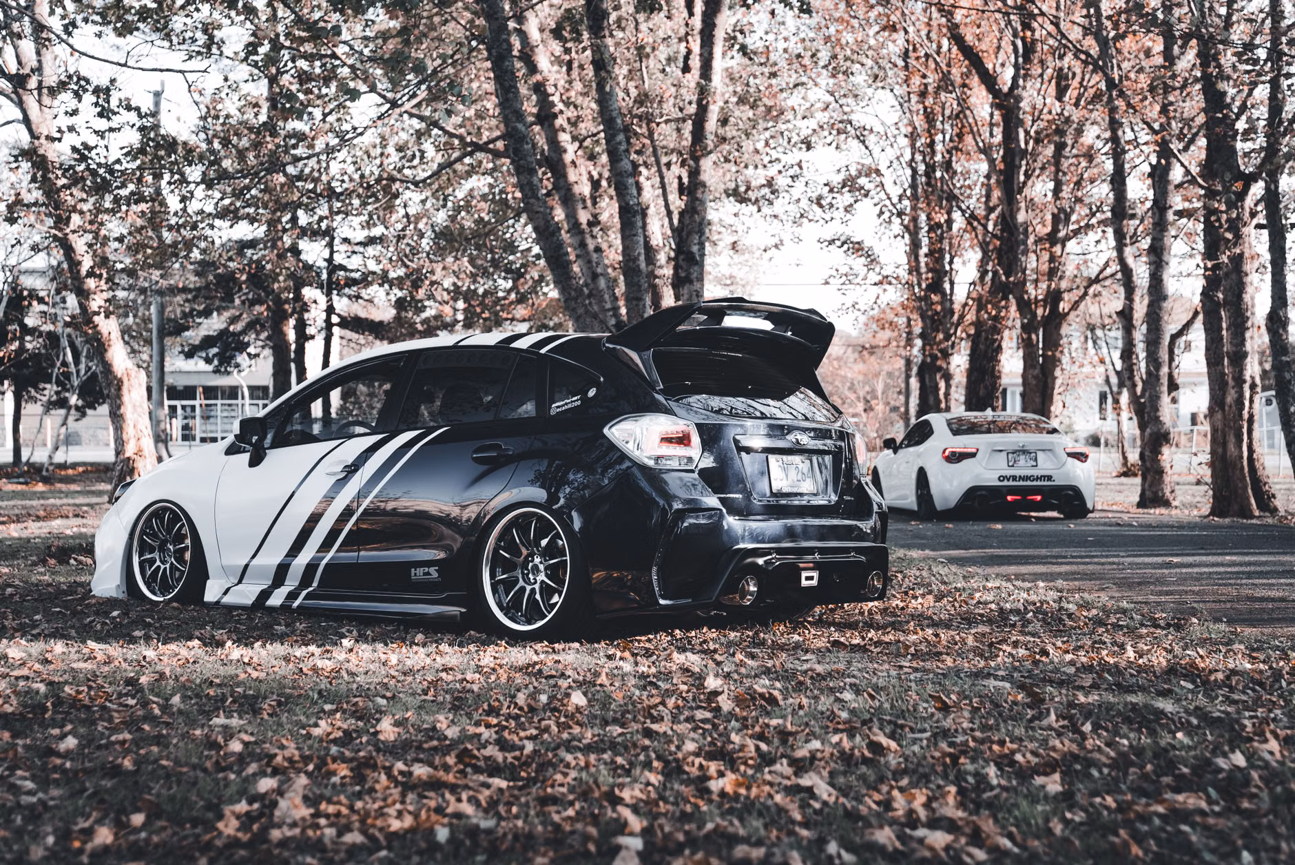 Due auto performanti personalizzate parcheggiate in un parco autunnale in una giornata di sole.